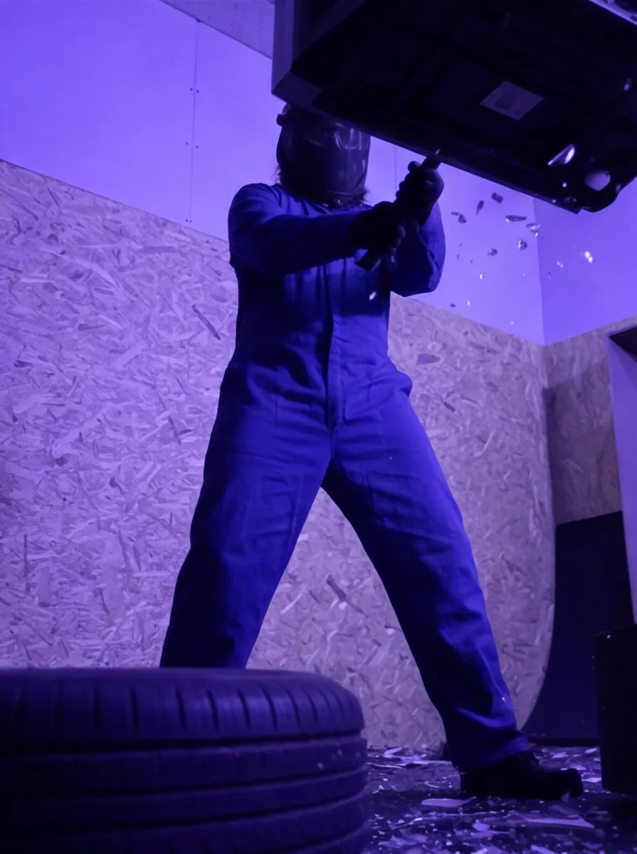 Customer in blue coveralls and safety mask smashing furniture with a sledgehammer in the Rage Room